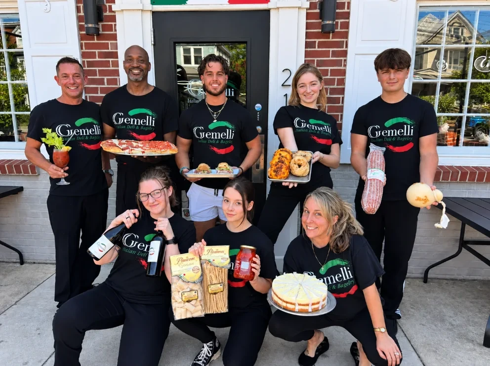 The Gemelli team in matching black Gemelli shirts outside the Pollock Street entrance, holding pizza, bagels, sandwiches, salami, provolone, wine, pasta and a cake