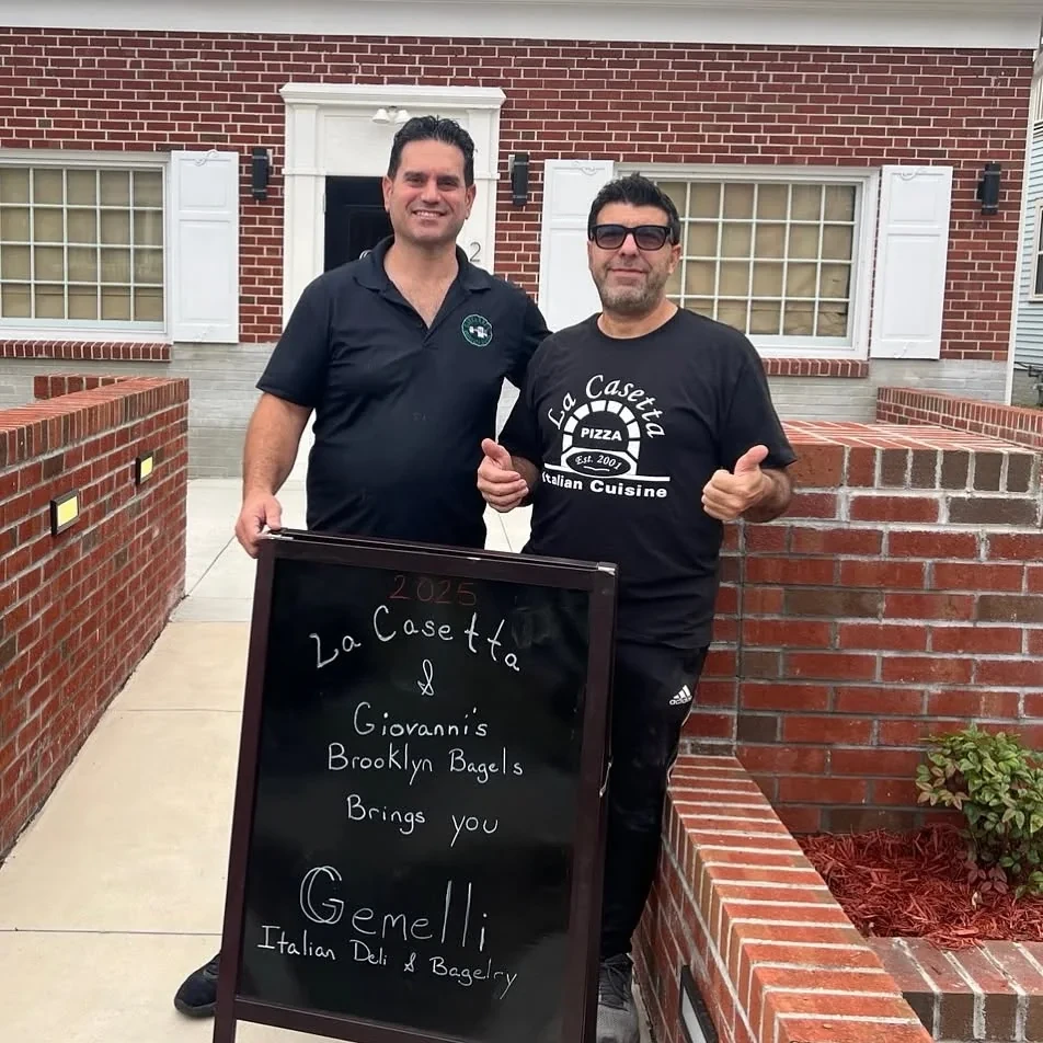 Giovanni Lentieri and Piero Passalacqua outside a brick building behind a chalkboard sign reading: La Casetta and Giovanni’s Brooklyn Bagels brings you Gemelli Italian Deli and Bagelry