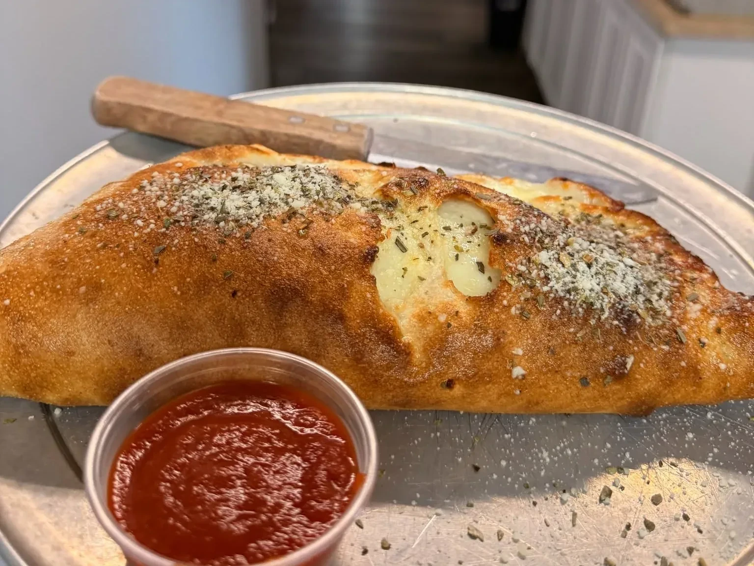A golden baked calzone on a tray with a cup of marinara for dipping